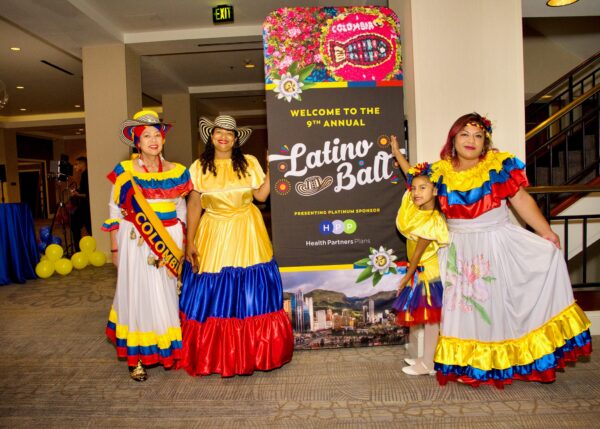 Three women and a young girl wearing colorful, flowy dresses, standing on the sides of a tall, Latino Ball narrow, retractable banner.